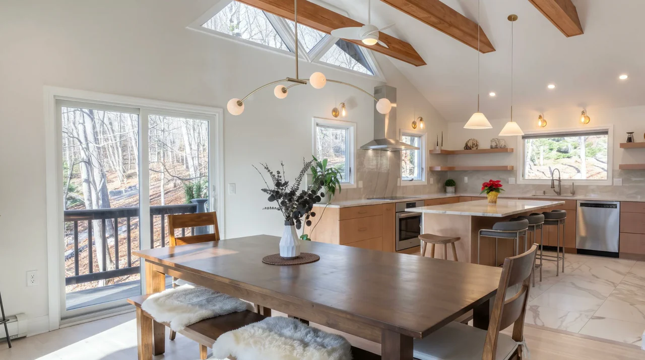 An open kitchen and dining room under a vaulted ceiling with wood beams, cherry cabinets, a marble island, brass globe lights and a wall of windows to the woods